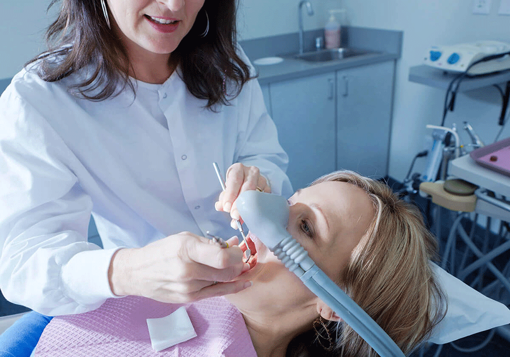 The image shows a dentist performing a dental procedure on a patient who is receiving nitrous oxide sedation through a nasal mask, with the dentist holding dental tools and working on the patient's mouth.