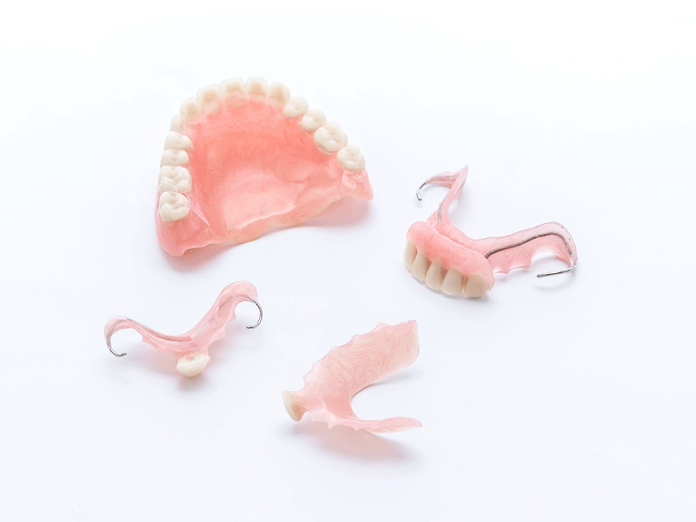 The image shows various types of dental prosthetics, including upper and lower dentures, as well as partial dentures with metal clasps, all placed on a white background.