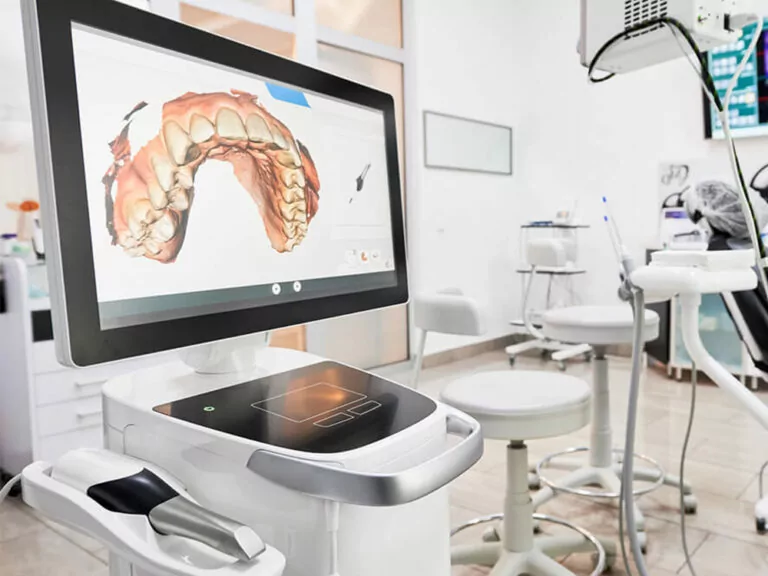 The image shows a modern dental office with a computer monitor displaying a 3D digital model of teeth, connected to a dental scanner, with dental chairs and instruments in the background, indicating a well-equipped clinic.