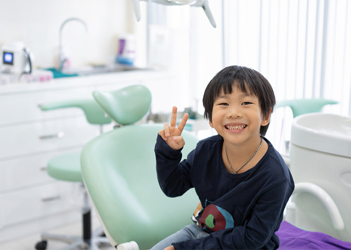 The image shows a young boy sitting in a dental chair, smiling and making a peace sign with his fingers, indicating a positive experience at the dentist's office.