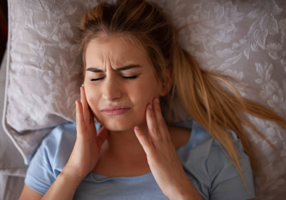 The image shows a woman lying in bed, holding her jaw with both hands and displaying a pained expression, indicating she is experiencing severe jaw pain or a toothache.