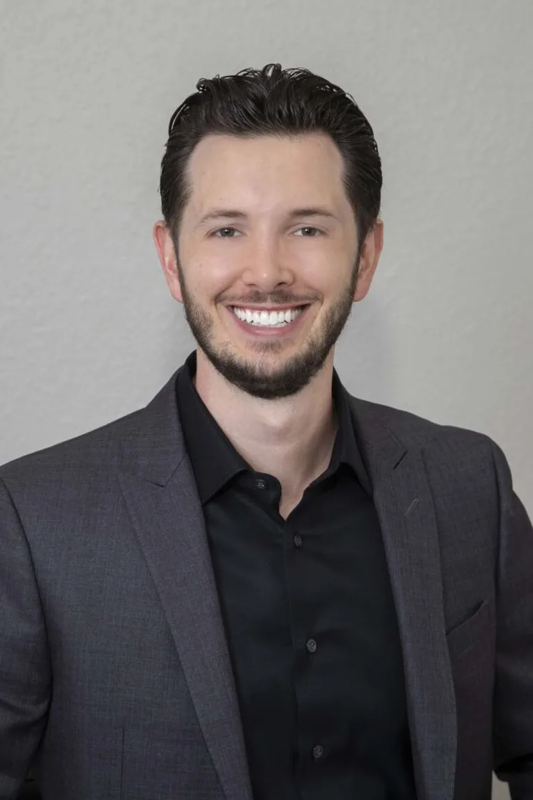 The image shows Dr. Jake Jacomet, a person wearing a dark grey suit jacket over a black dress shirt, with short dark hair and a neatly trimmed beard. The background is a plain, light-colored wall.