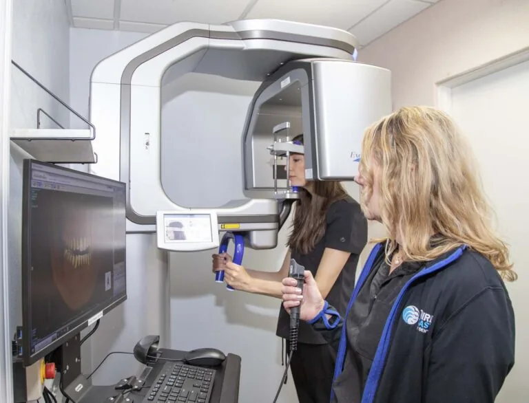 The image shows a CBCT (Cone Beam Computed Tomography) machine, used for capturing detailed 3D images of a patient's teeth, jaw, and surrounding structures. A dental professional operates the machine while another screen displays the captured dental images.