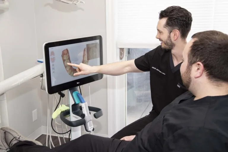 Dr. Jacomet in a black embroidered shirt points at a screen displaying a 3D model of teeth, guiding a seated patient. The screen includes icons related to dental imaging, reflecting advanced technology in dental care.