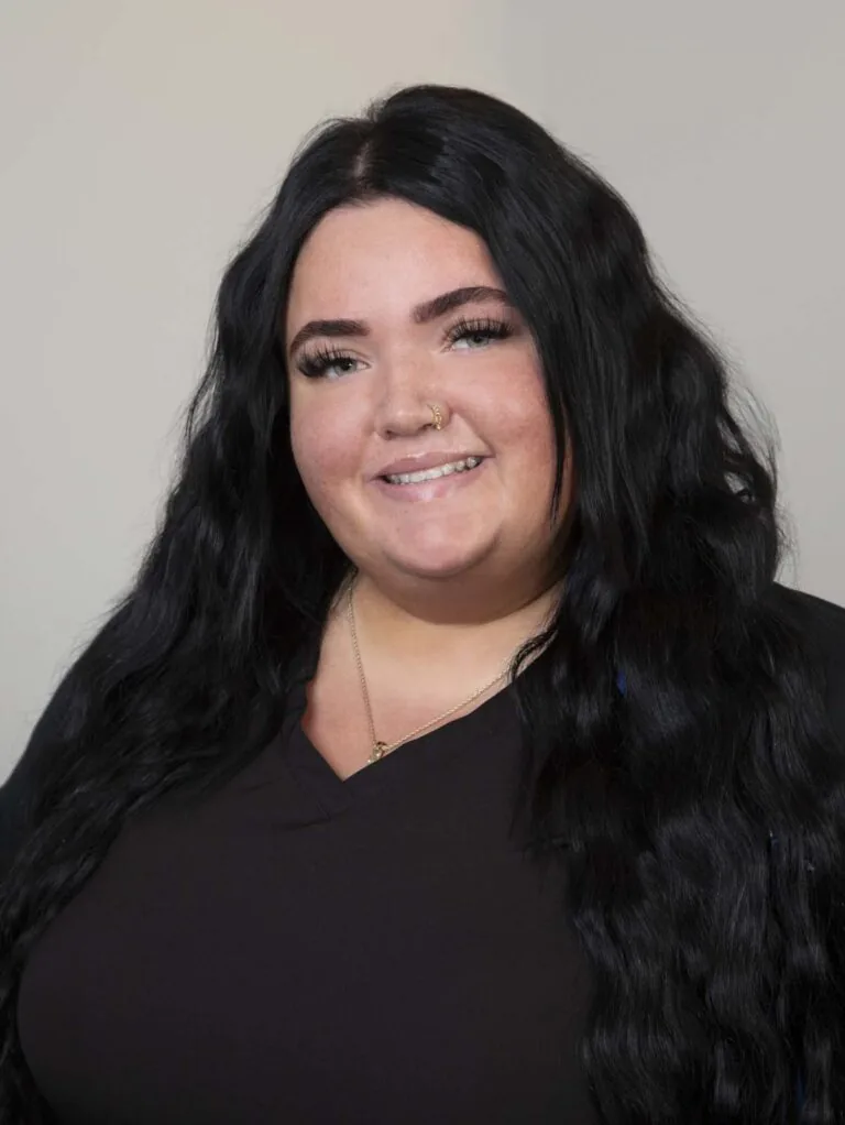 The image depicts Courtney Stout, a person with long, wavy black hair, wearing a dark top and necklace, against a plain background.