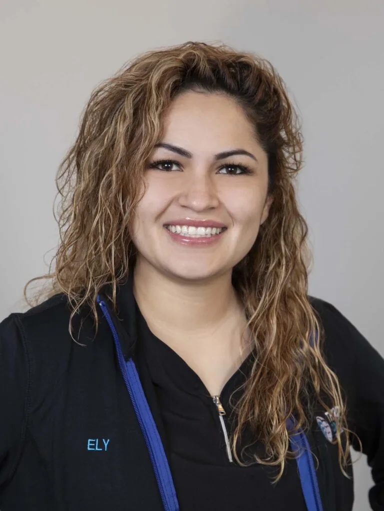 The image shows Elizabeth (Ely) Perez, a person with long, wavy hair wearing a black jacket with blue accents and a black shirt underneath. The jacket has the name "ELY" embroidered on it.