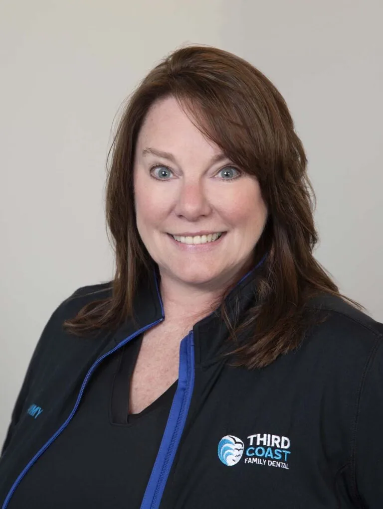The image shows a woman smiling, wearing a black jacket with blue trim and the name "TAMMY" embroidered on the left side. The logo "THIRD COAST FAMILY DENTAL" is visible on the right side of the jacket.